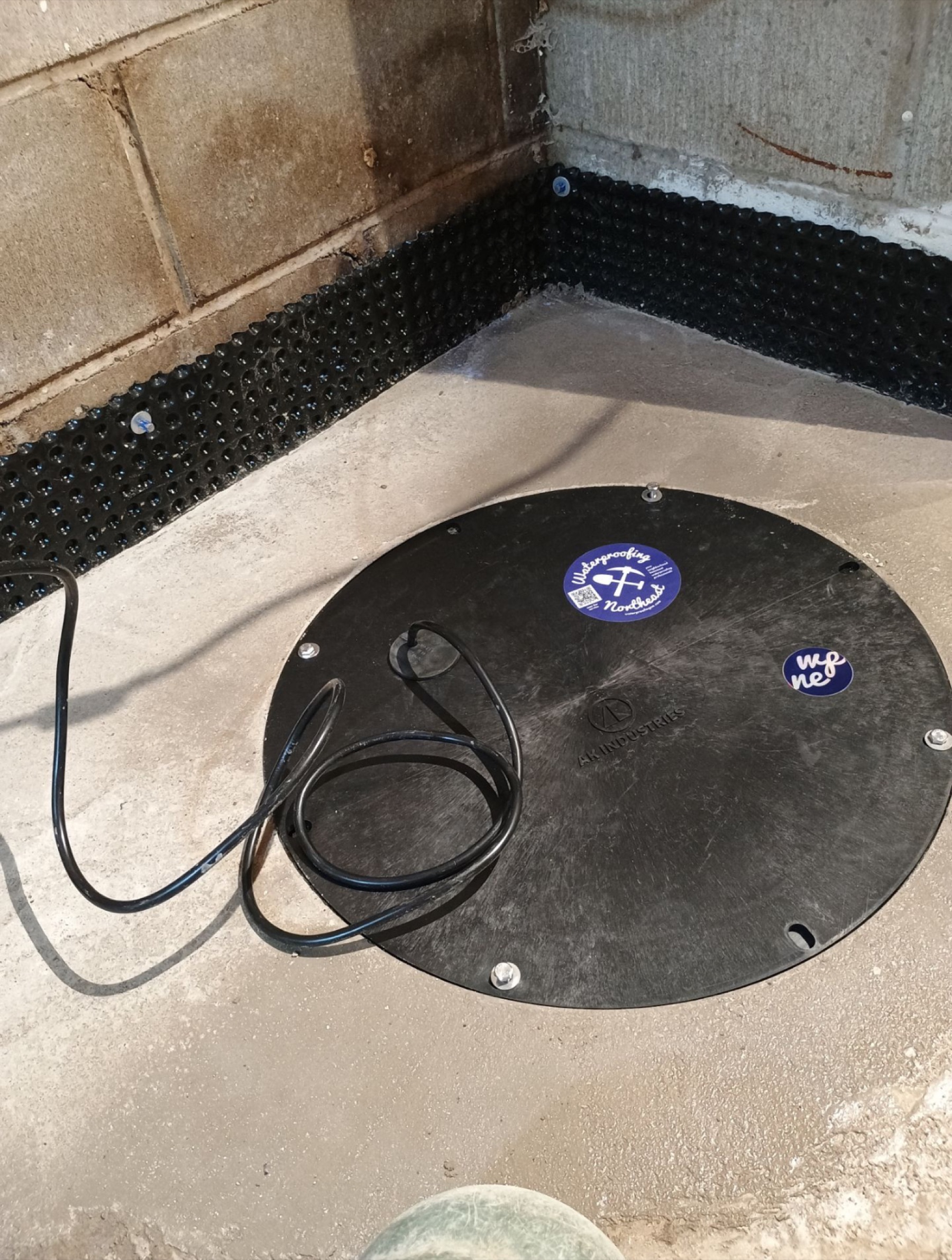 A finished sump basin lid with the Waterproofing Northeast company sticker, set in fresh concrete beside the dimple-membrane wall of an interior drain tile install